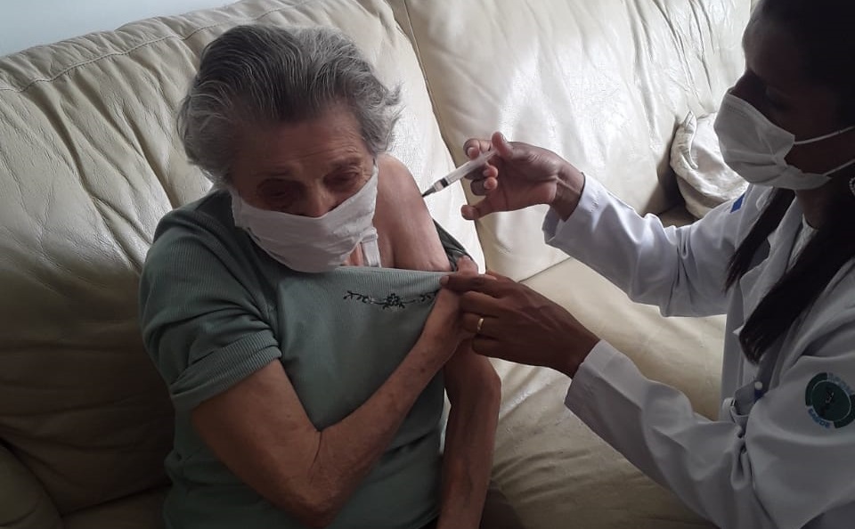 Foto de vacinação. Uma idosa está sentada em um sofá marrom claro. Ela tem cabelos grisalhos, veste uma blusa de frio verde claro e está abaixando uma das mangas para receber a dose da vacina de reforço, aplicada por uma profissional de saúde que está sentada ao seu lado.