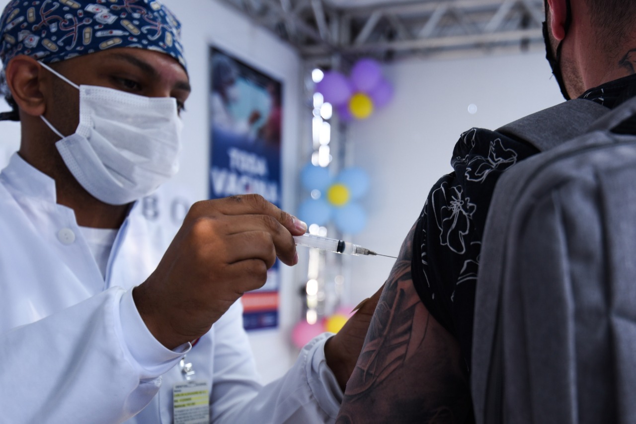 À esquerda da imagem, um profissional de saúde vestindo jaleco branco, máscara branca e uma touca azul na cabeça está aplicando a vacina no braço de um homem que está de costas. Ele usa uma camisa preta com estampa branca, tem tatuagem no braço e está com uma mochila cinza nas costas.