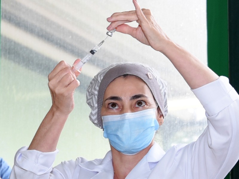 Profissional da saúde prepara uma dose da vacina para ser aplicada. Ela segura a seringa numa das mãos e o frasco da vacina na outra