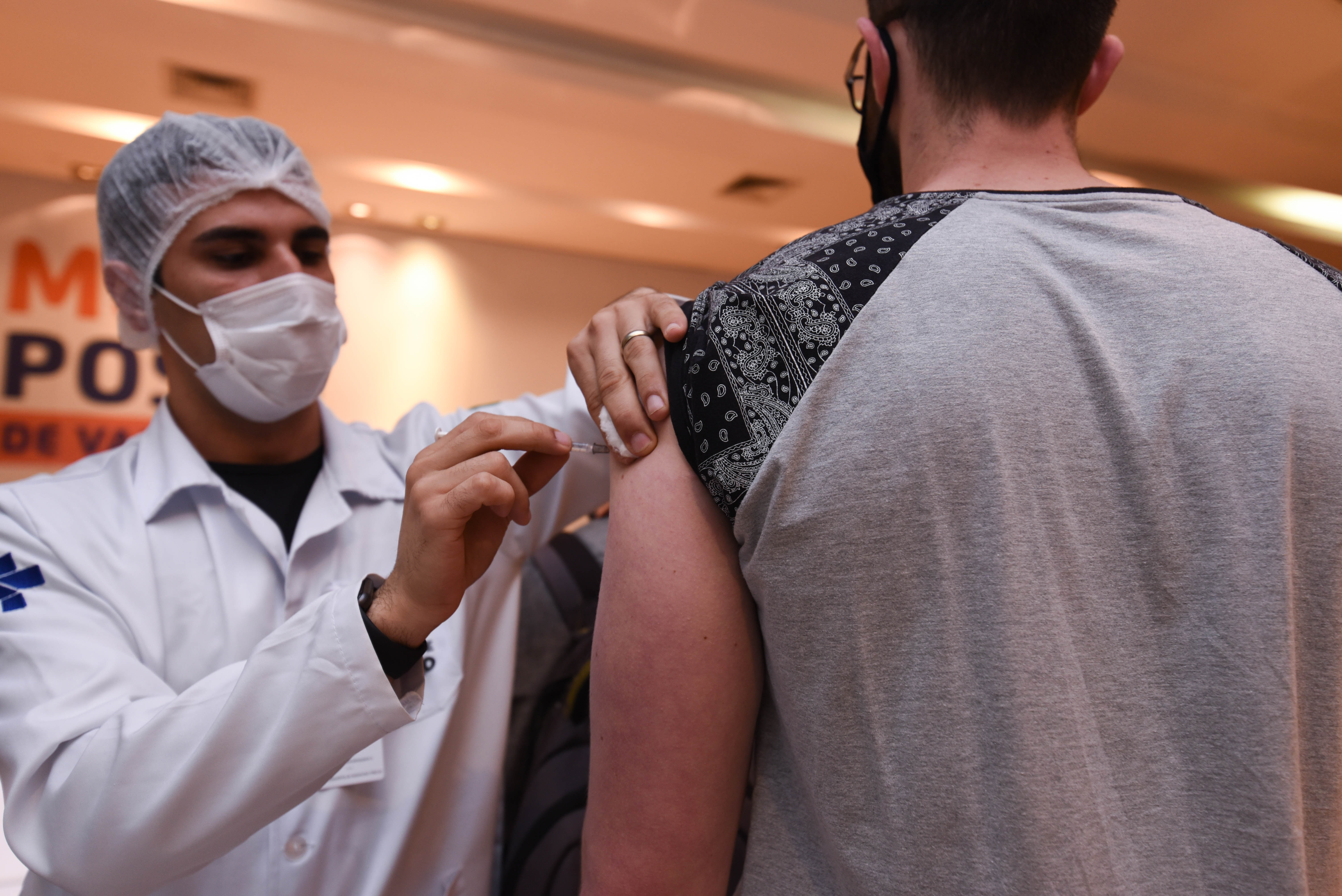 Foto de vacinação. Um profissional de saúde vestindo jaleco, máscara e touca aplicando vacina no braço de homem que está em pé virado de costas vestindo camiseta cinza com a manga levantada.