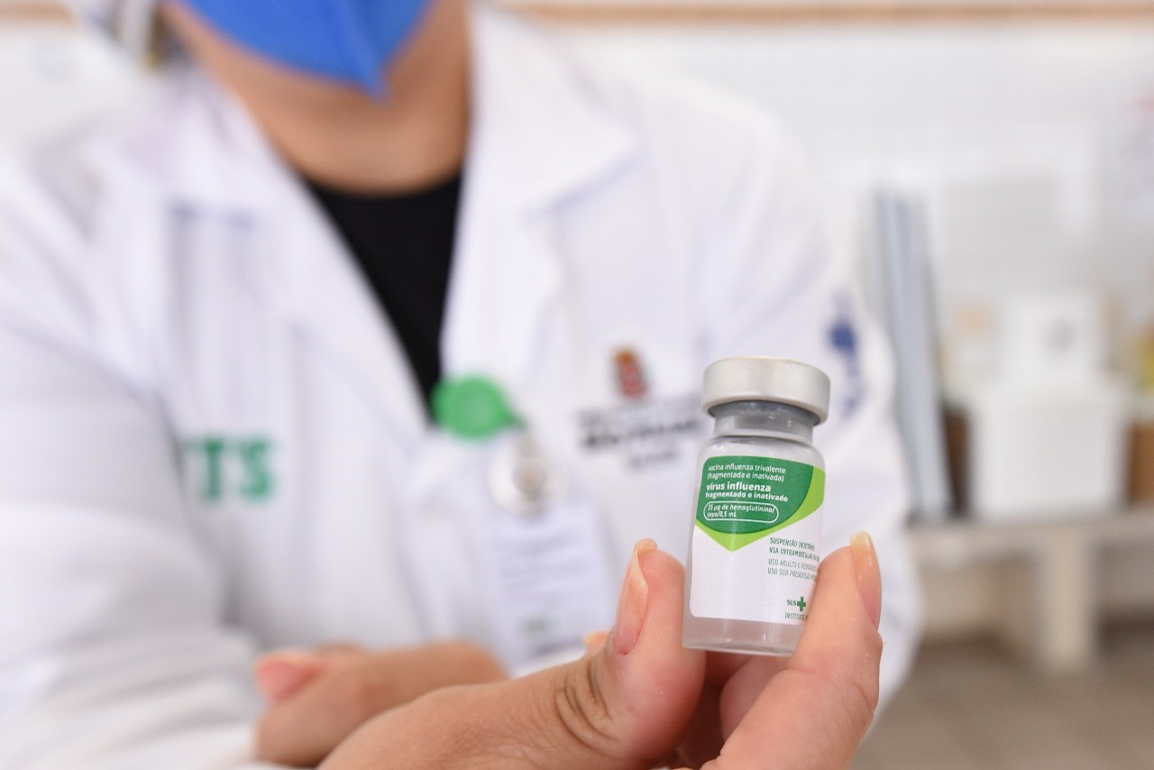 Foto de frasco de vacina. A mão de uma profissional de saúde vestindo um jaleco branco está segurando um frasco de vacina transparente, com o rótulo branco e verde e tampa cinza. No rótulo está a frase “Vírus influenza fragmentado e inativado”.