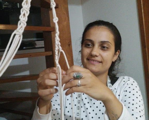 Uma mulher vestindo uma blusa de mangas longas branca com estampa de bolinhas pretas está tecendo cordas, que estão penduradas em um suporte no alto.
