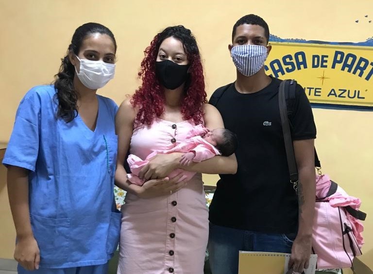 Uma profissional de saúde vestindo uniforme azul está usando máscara branca de proteção. Ela posa ao lado de um casal. A mulher tem cabelos rosas, está usando máscara de proteção preta e segura um recém-nascido no colo. Ao lado dela, está um homem de máscara de proteção listrada e camisa preta.