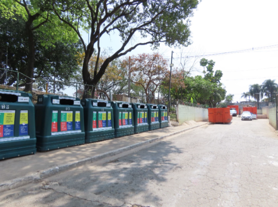 Foto de um Ecoponto, uma área destinada à coleta seletiva de diversos tipos de resíduos. No primeiro plano, à esquerda, há vários contêineres grandes, verdes e pretos, com aberturas e indicações de materiais recicláveis. Alguns paletes de madeira estão encostados nos contêineres. Ao fundo, à direita, três grandes contêineres laranjas, do tipo caçamba estacionária, estão vazios e abertos. O chão da área é de terra batida e cascalho. Árvores com folhagem verde cercam o local, e alguns edifícios residenciais de cores claras são visíveis ao fundo, sob um céu azul com algumas nuvens brancas. A luz do sol ilumina a cena.