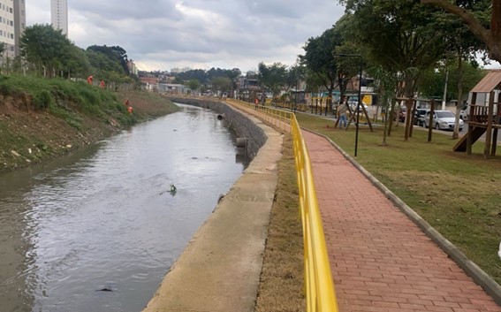 Foto de um canal de rio com água escura e turva, correndo em paralelo a um caminho de tijolos vermelhos. À direita, o caminho é delimitado por uma grade amarela e grama verde. No lado esquerdo, o rio é cercado por um muro de concreto cinza. Ao fundo, árvores e prédios residenciais são visíveis sob um céu nublado. A foto foi tirada em um ângulo que mostra a extensão do canal e do caminho, sugerindo um espaço urbano de lazer ou infraestrutura.