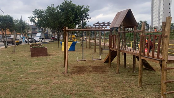  Foto de um parquinho infantil em um dia nublado. No centro, há um balanço de madeira com duas cadeiras penduradas. À direita, um playground de madeira com escorregador e escada é visível. À esquerda, um canteiro de madeira quadrado com plantas está no chão. Ao fundo, uma rua com carros e edifícios residenciais pode ser vista. O chão do parquinho é gramado e há árvores espalhadas pela área.