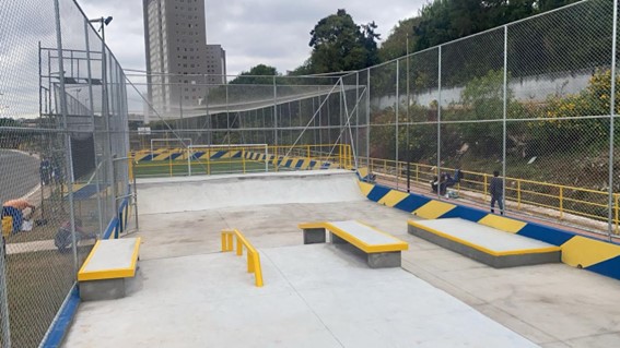 Foto de uma pista de skate recém-construída, vista de cima e em perspectiva. O piso da pista é de concreto cinza claro, com bancos de concreto e metal amarelo dispostos em diferentes pontos. Ao fundo, uma cerca de tela metálica delimita a área, com árvores e um prédio alto visíveis além da cerca. A pista inclui obstáculos como rampas e corrimãos, e há uma área gramada com uma pequena trave de gol no canto superior esquerdo. Duas pessoas, vestindo roupas escuras, estão em pé na lateral da pista, à direita. O céu está nublado, com tons de cinza e branco.