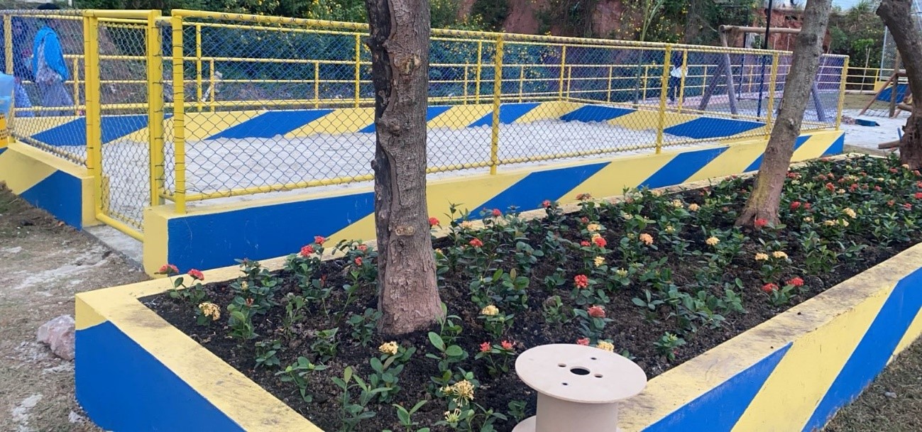 Foto de um parque com um espaço cercado por grades amarelas e azuis, com um padrão listrado diagonal. Dentro do cercado, há um piso de concreto cinza claro. No primeiro plano, um canteiro retangular com flores vermelhas e amarelas está em frente ao cercado. Árvores finas e altas crescem entre o cercado e o canteiro. Ao fundo, mais árvores e parte de um playground de madeira são visíveis. No canto inferior direito, um objeto redondo de madeira, possivelmente um carretel, está no chão. O dia parece ensolarado e a luz destaca as cores vibrantes do cercado e das flores.