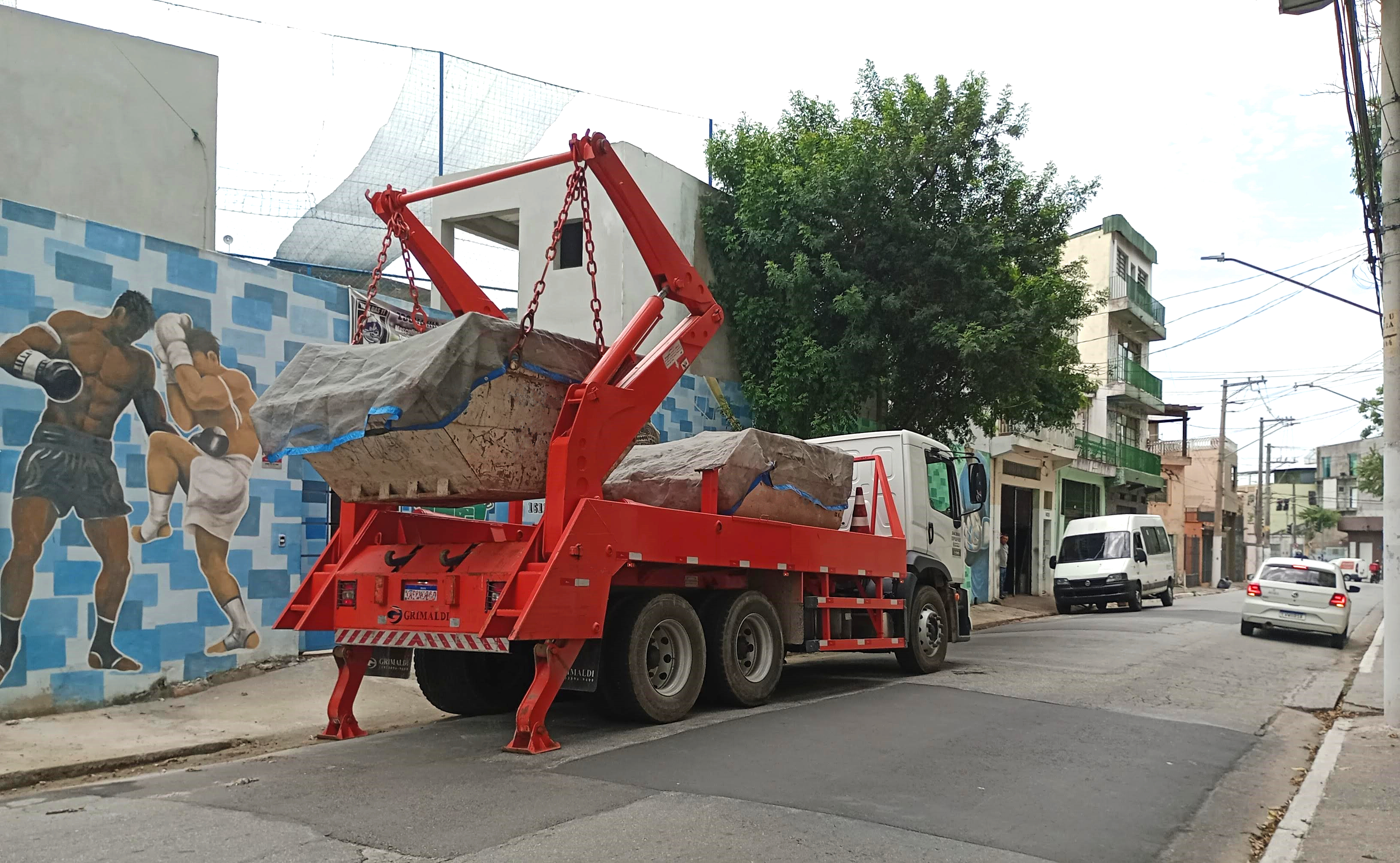 Um caminhão poliguindaste vermelho realiza o transporte de caçambas cobertas por lonas em uma rua urbana. Ao fundo, um mural artístico com uma cena de boxe decora a parede de um prédio. O veículo está estacionado próximo à calçada, realizando o processo de carga ou descarga das caçambas, enquanto outros veículos circulam pela via. A cena indica uma operação de coleta de resíduos ou entulho, possivelmente relacionada à fiscalização ambiental ou gestão de resíduos sólidos.