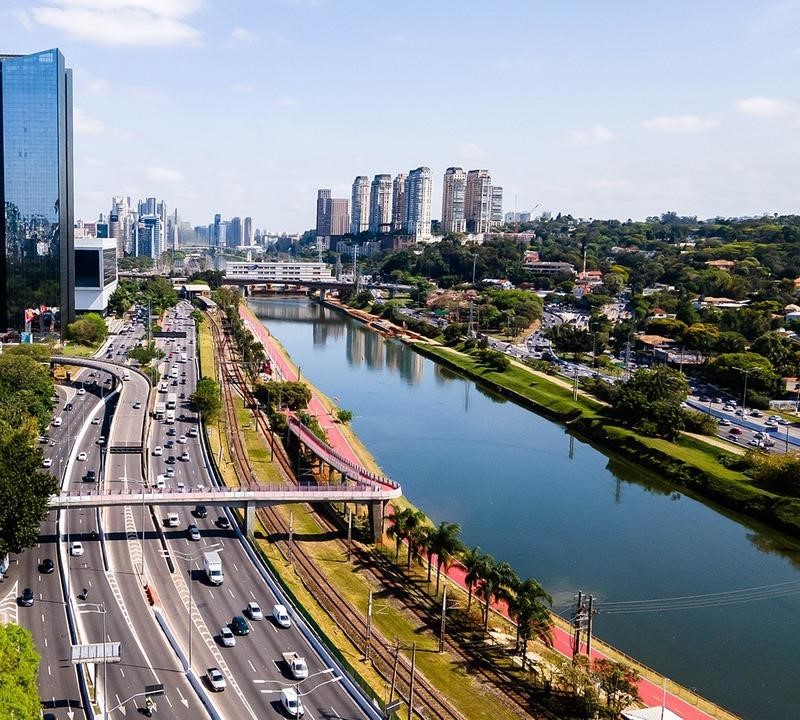 A imagem mostra uma paisagem urbana de São Paulo, Brasil, destacando a Marginal Pinheiros com suas múltiplas faixas de tráfego, ciclovia e o Rio Pinheiros ao centro. À esquerda, há um grande edifício envidraçado refletindo o céu, enquanto ao fundo se observa um conjunto de prédios altos em uma área nobre da cidade. A cena também inclui passarelas para pedestres, árvores ao longo das vias e áreas verdes ao redor, criando um contraste entre a urbanização e a natureza. O céu está azul com poucas nuvens, e o trânsito na via expressa está fluindo.
