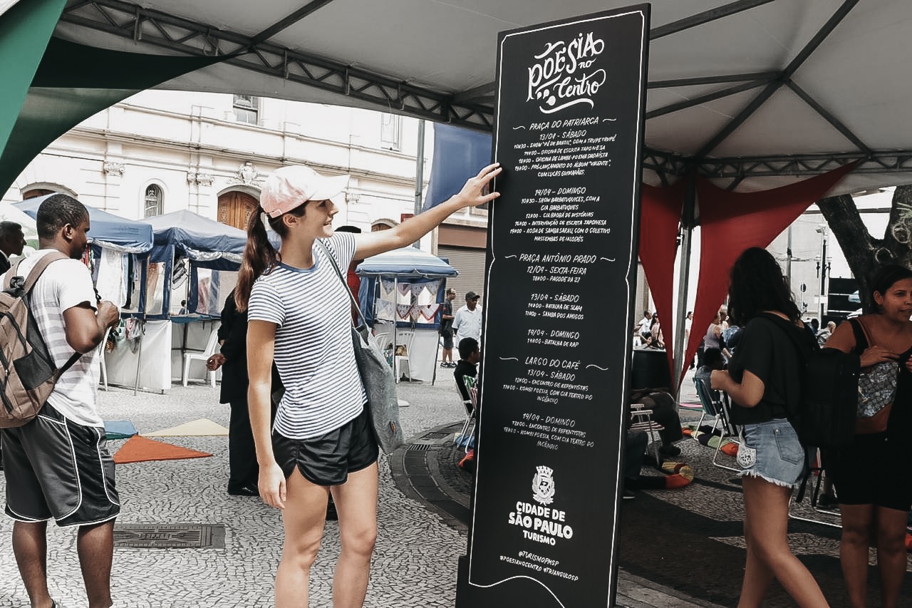 Foto de uma jovem com a mão na placa Poesia no Centro lendo os locais dos eventos.