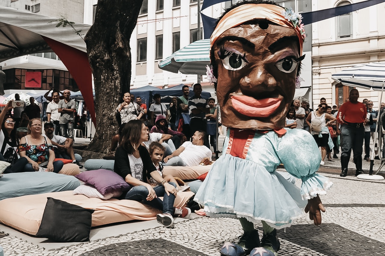 Foto de um boneco gigante de uma baiana com várias pessoas sentadas em almofadões assistindo o evento.
