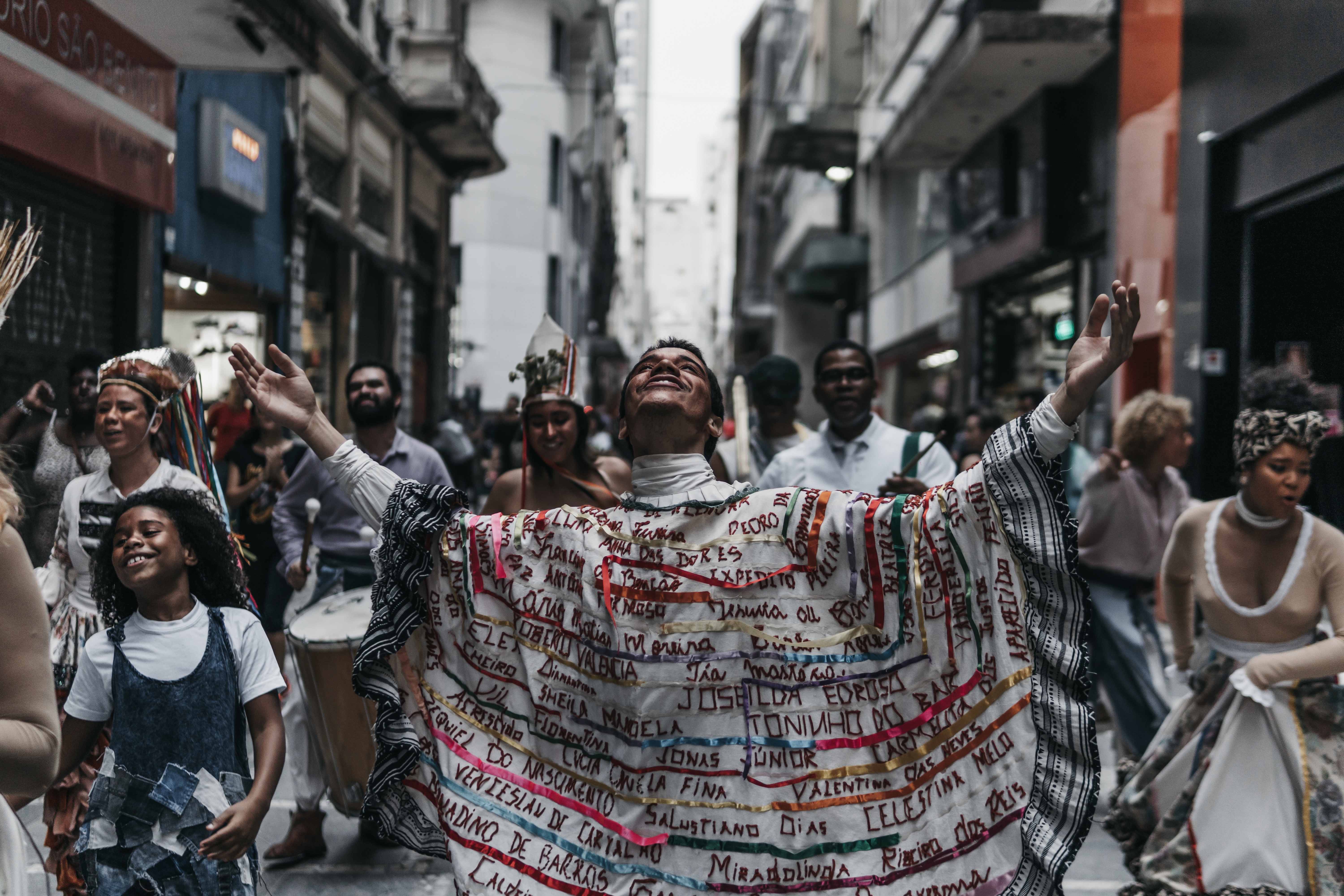 Foto de um jovem com uma camisa com várias fitas coloridas coladas e o nome de diversas pessoas bordado. Em volta dele tem várias pessoas fantasiadas e algumas com instrumentos musicais.