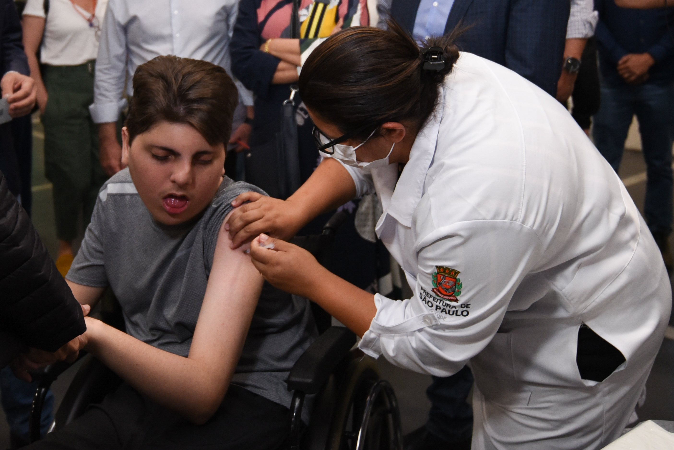 Foto de vacinação. Profissional da saúde usando máscara e jaleco branco com simbolo da prefeitura de São Paulo bordado na manga aplica vacina no braço de um adolescente com deficiência que está em cadeira de rodas. O menino usa camiseta cinza com uma das mangas levantadas. Ao fundo é possível ver algumas pessoas em pé.