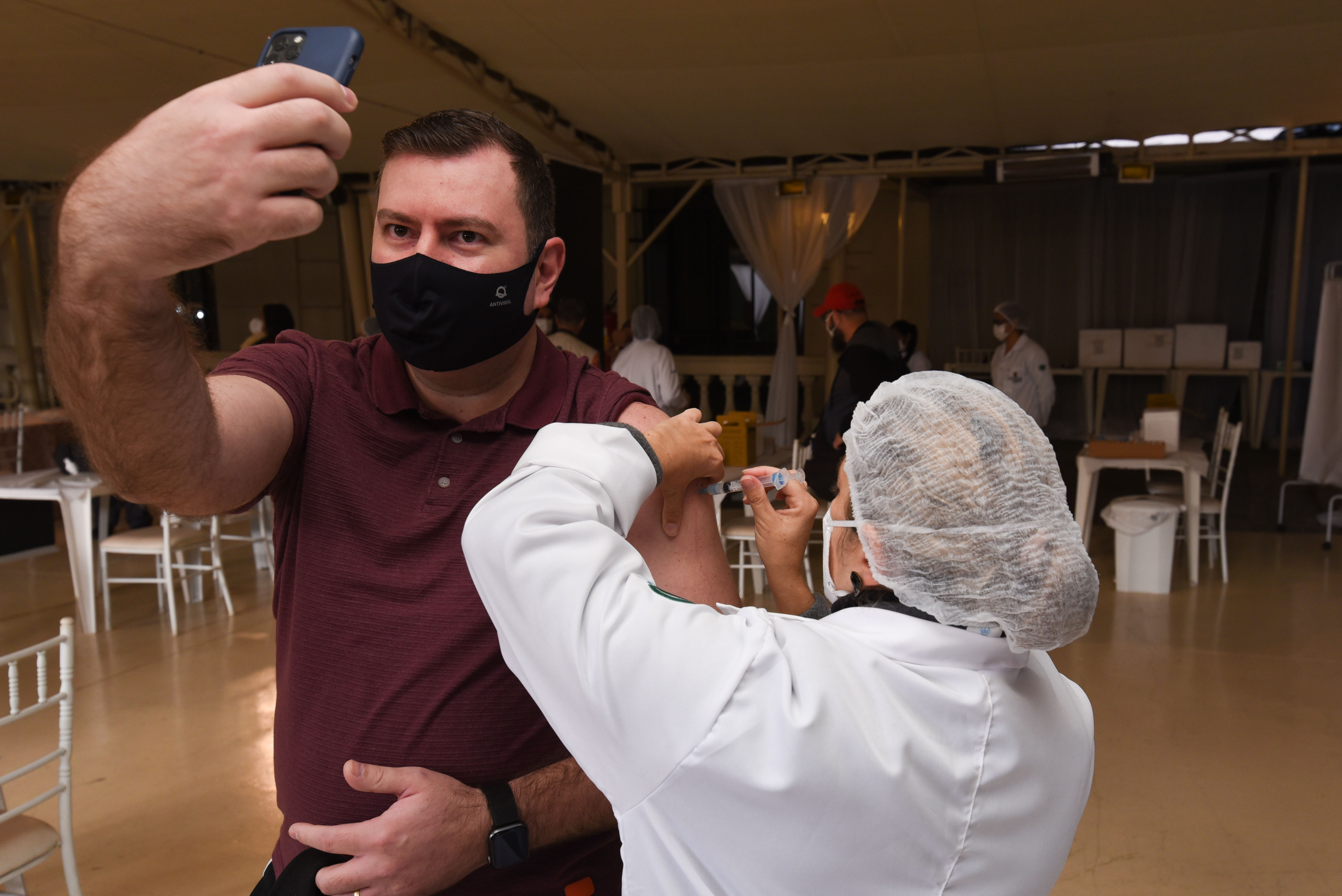 Foto de vacinação. Homem de máscara preta e camiseta segura o celular para tirar foto enquanto uma profissional da saúde vestida de jaleco, touca e máscara aplica a vacina nele. Ao fundo é possível ver algumas mesas e cadeiras e outros profissionais da saúde.
