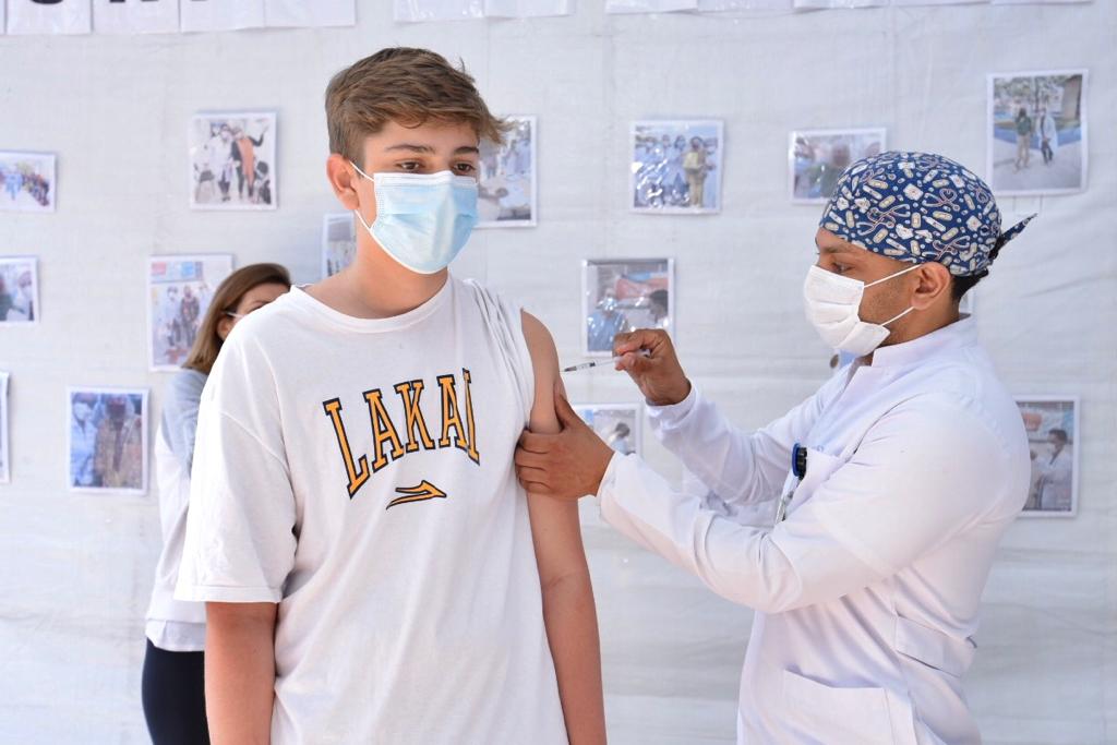 Foto de vacinação. Um profissional de saúde vestindo jaleco branco, máscara de proteção branca e uma touca azul com detalhes em branco na cabeça está aplicando a dose da vacina em um jovem mais alto. Ele veste uma camisa branca com detalhes em amarelo.