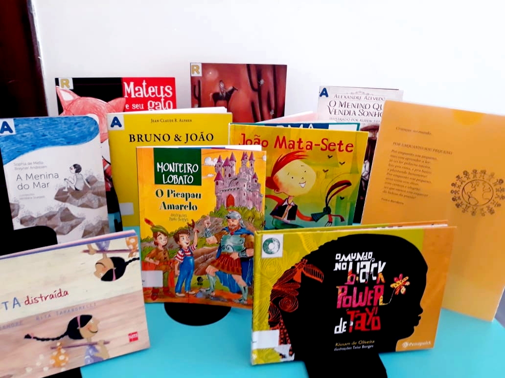 Foto de nove livros sobre superfície azul. As capas tem tons de amarelo, laranja, vermelho, branco e azul.