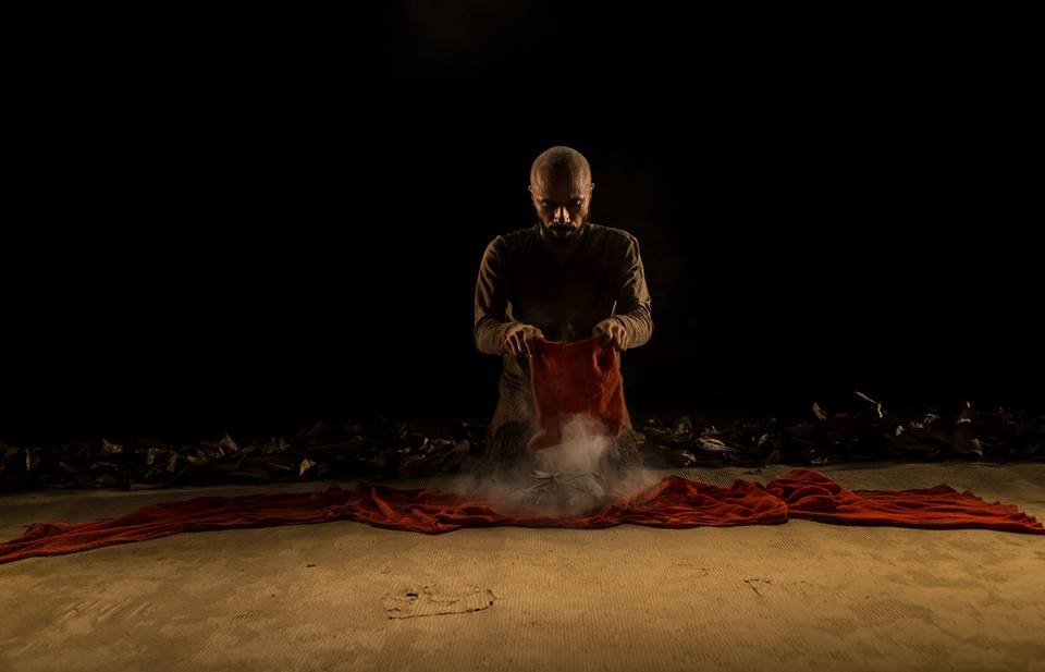 Foto em tons escuros de homem ajoelhado segurando um pano vermelho sobre o chão, da onde sai fumaça