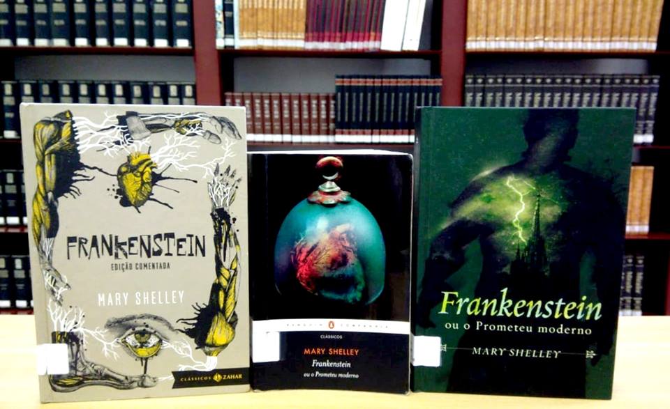 Foto de três edições do livro Frankenstein sobre uma superfície de cor creme, ao fundo, estantes com livros. Da esquerda para direita, o primeiro livro tem capa cinza e em seu entorno há ilustrações de músculos de braços, pé, olho, veias e um coração na parte superior, na sequência, o título em letras estilizadas na cor preta. Mais abaixo, próximo à imagem do olho, o nome da autora na cor branca. O segundo livro tem fundo preto com ilustração de um coração dentro de uma redoma de vidro azul, na parte inferior está o nome da autora na cor laranja e, na sequência, o título na cor branca. A última edição tem capa verde com ilustração de uma sombra masculina, dentro dela há imagem de um castelo e raios verdes. O título está na parte inferior em tons de verde e branco, abaixo, o nome da autora na cor bege.