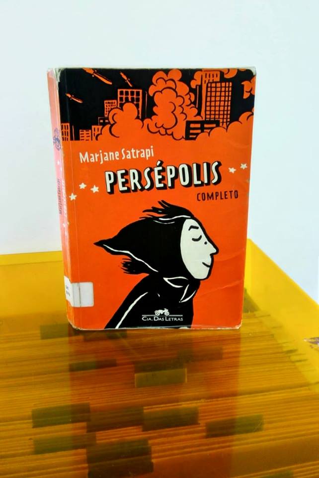 Livro sobre superfície laranja, ao fundo, parede branca. Capa do livro de fundo laranja, com desenho de um bombardeio e prédios na parte superior. No meio, o título e o nome da autora, escritos na cor branca e abaixo, ilustração em preto e branco de uma garota com hijab ao vento.