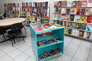 Interior da biblioteca com estandes e caixas expositoras de livros, além de mesas e cadeiras