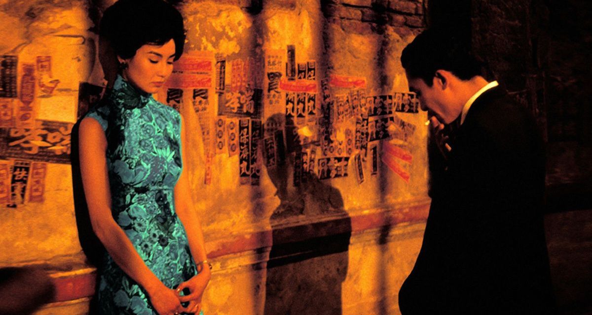 Foto apresenta um casal cabisbaixo em frente à uma parede colorida. Ela veste um vestido em tom verde; Já o homem aparece fumando e utiliza um terno de cor preta.