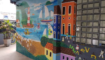 Parede grafitada com desenho depraia, com um farol, barcos a vela, pessoas de roupas brancas na areia e casinhas coloridas