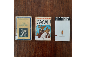 foto de 3 livros sobre a mesa vistos de cima: "Dom Casmurro", de Machado de Assis; "Cacau", de Jorge Amado; e "A obscena senhora D.", de Hilda Hilst