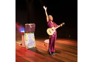 Mulher no palco, com roupas roxas, com um violão pendurado no ombro, com o braço direito suspendido