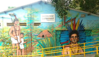 Fachada de uma casa decorada com uma pintura mostrando tres indigenas e uma oca, em meio a vegetacao e ceu azul