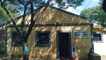 Fachada de uma casinha amarela com duas janelas quadradas e uma porta, todas emolduradas por grossas faixas cinzas. Em frente, uma arvore cria sombras pontilhadas na construcao.