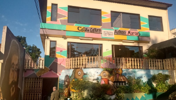 Casa de tres andares com paredes decoradas com formas geometricas em tons de amarelo, verde, rosa e marrom. Na parte terrea, um muro se destaca com desenhos de passaros