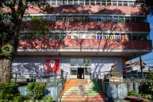 Fachada mostrando tres andares de um predio, sombreados por arvores altas. Uma escada decorada leva ao portao de entrada