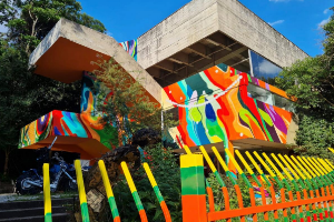 Edificio em concreto armado, com a metade inferior pintada em formas organicas de diversas cores vivas. Ceu azul ao fundo e arvores dos dois lados