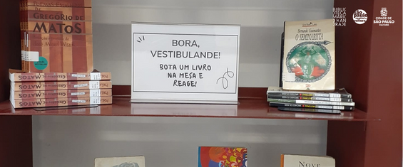 foto de prateleira em que se vê, entre duas pilhas de livros, uma placa com os seguintes escritos: "Bora, vestibulande! Bota um lvro na mesa e reage"