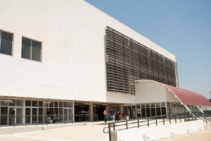 Fachada de um edificio branco com o terreo formado por janelas de vidro e pilares. Ceu azul ao fundo