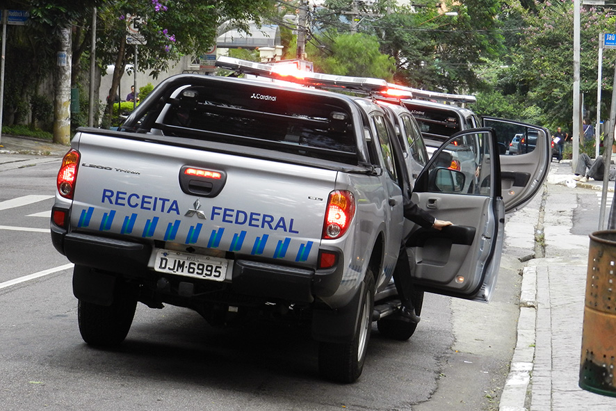 Foto mostra a parte traseira de três veículos da Receita Federal, do tipo viatura, com as portas abertas e giroflex ligado. No veículo em primeiro plano, vemos o braço de uma pessoa abrindo a porta