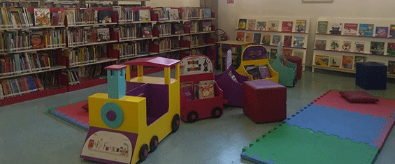 Interior da Biblioteca Sylvia Orthof com as estantes com livros, um trenzinho com livros infantis e pufes para sentar
