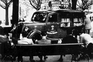 Ford caminhonete-biblioteca do Departamento da Cultura por volta de 1940 parado numa praça com leitores sentados num banco do jardim.