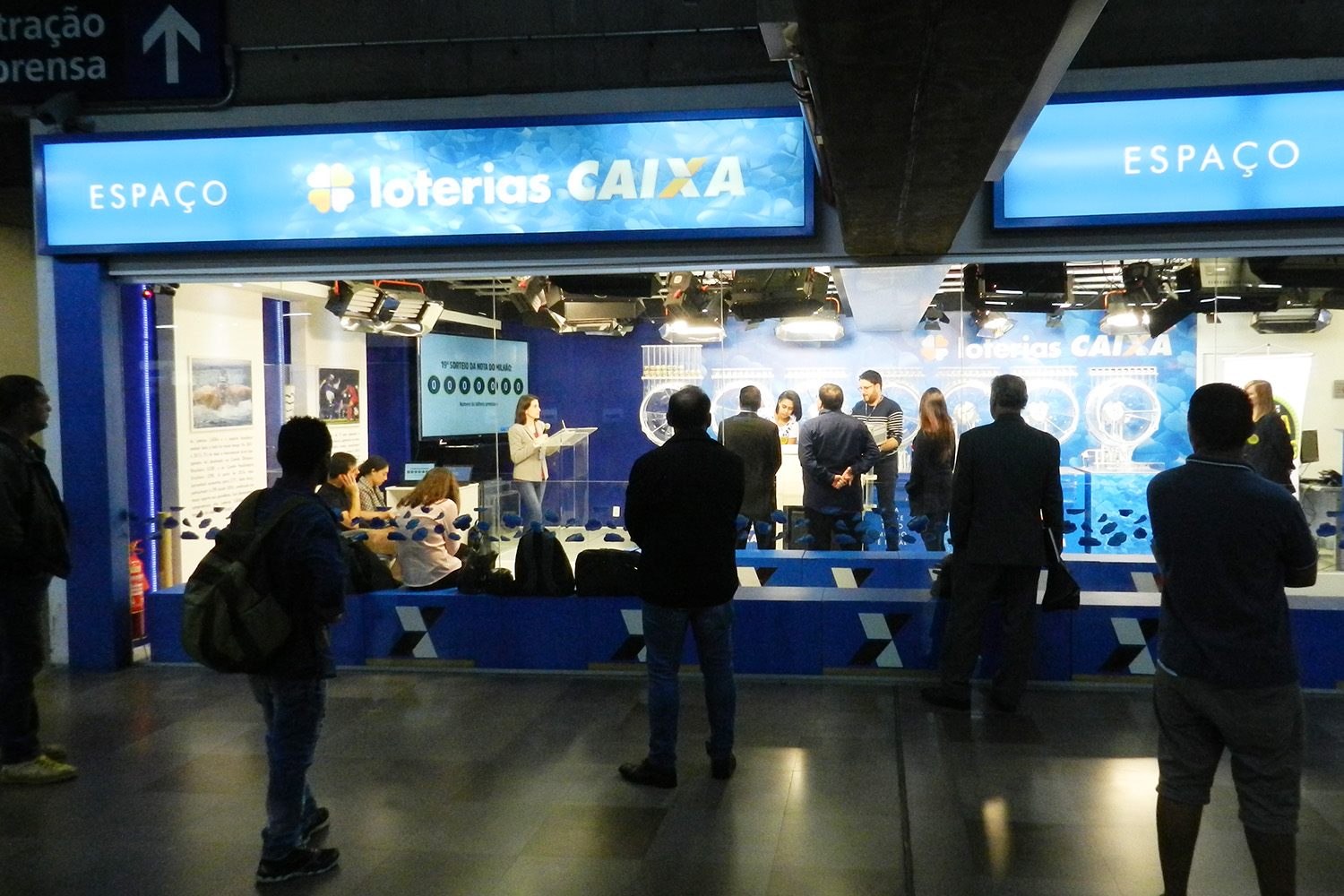 Imagem mostra a fachada do Espaço Loterias Caixa, no Terminal Tietê, com letreiro azul na parte de cima e paredes de vidro. Cinco pessoas de costas observam, de fora do espaço, o sorteio que é realizado do lado de dentro.