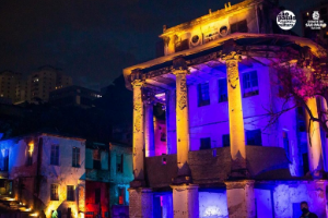 Palacete do início do século XX com pilares altos e uma estrutura antiga preservada, ao lado de casinhas geminadas, formando uma vila. Toda a construção é iluminada em tons de azul, rosa e amarelo, com um detalhe em vermelho ao canto. É noite.