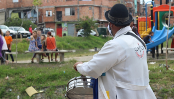 Um homem esta de costas tocando uma caixa e olhando para as pessoas que se divertem numa praça