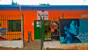 Casa terrea com parede vermelha e listra azul no topo, com pinturas mostrando o rosto de uma mulher negra, a frente, e o busto de um homem negro ao fundo