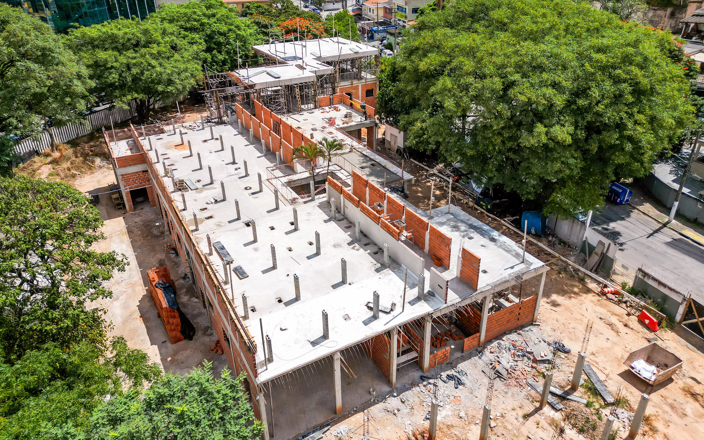 A imagem mostra uma obra em andamento vista de cima, provavelmente registrada por drone. Trata-se de um prédio de médio porte em construção, com estrutura de concreto já concluída em parte e paredes de tijolos aparentes sendo erguidas. É possível ver lajes prontas com diversos pilares verticais expostos, indicando que o edifício terá mais de um pavimento. Em algumas áreas, há andaimes montados e trabalhadores atuando, sugerindo que a obra está em fase estrutural e de alvenaria. O entorno é bastante arborizado, com árvores grandes ocupando boa parte do terreno ao lado e criando contraste com a construção. Também há uma rua próxima, com calçada, postes e alguns equipamentos urbanos visíveis. No chão, ao redor da obra, aparecem materiais de construção espalhados, como tijolos, entulho e caçambas, além de áreas de terra batida.