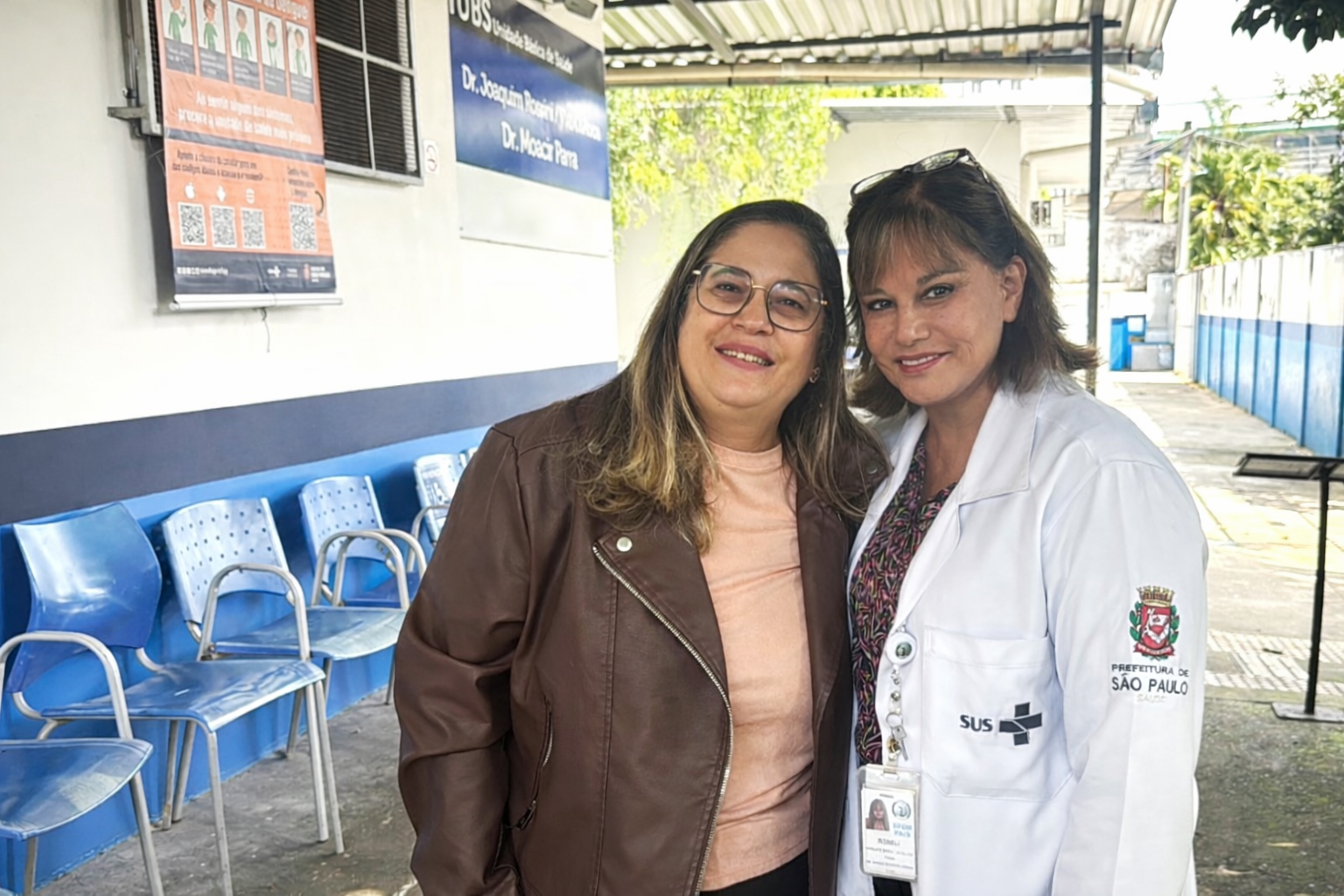 A imagem mostra Simone Vigilato, participante assídua do Avança Saúde vestindo uma blusa em tom claro e uma jaqueta marrom. Ela sorri de forma acolhedora, com postura próxima e confiante, demonstrando vínculo com o serviço. Ao seu lado, está a gerente da UBS Joaquim Rossini, usando jaleco branco com identificação da Prefeitura de São Paulo e do SUS. Ela também sorri para a câmera, transmitindo segurança e profissionalismo.