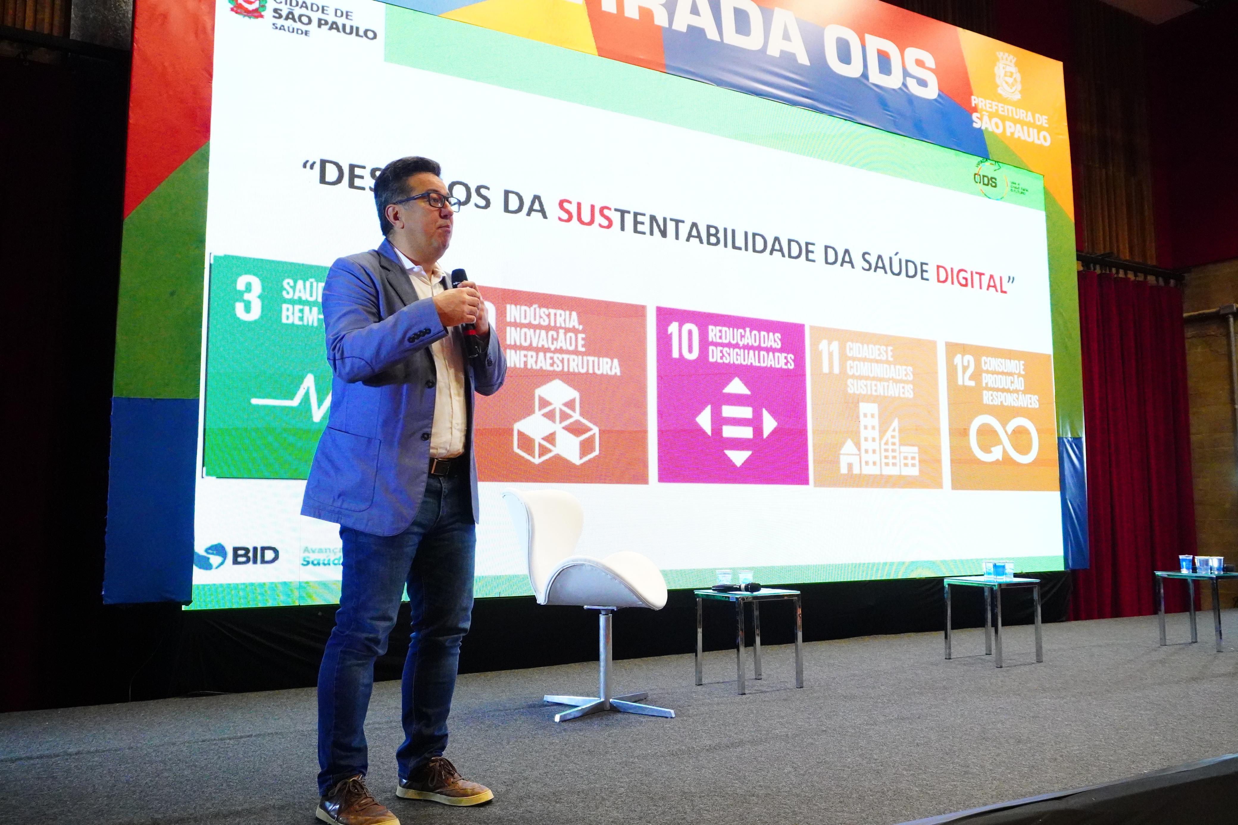 A imagem mostra um homem palestrando em um evento da Prefeitura de São Paulo sobre os Objetivos de Desenvolvimento Sustentável (ODS). Ele está de pé no palco, segurando um microfone, com um telão ao fundo que exibe o título da apresentação: “Desafios da Sustentabilidade da Saúde Digital”. No painel também aparecem cinco ODS relacionados ao tema: ODS 3: Saúde e bem-estar ODS 9: Indústria, inovação e infraestrutura ODS 10: Redução das desigualdades ODS 11: Cidades e comunidades sustentáveis ODS 12: Consumo e produção responsáveis O ambiente é formal, com mesas e cadeiras no palco, e o evento parece focado em inovação e políticas públicas de saúde. No canto superior direito, aparece o logotipo da Prefeitura de São Paulo – Saúde, e no canto inferior esquerdo, o do BID (Banco Interamericano de Desenvolvimento).