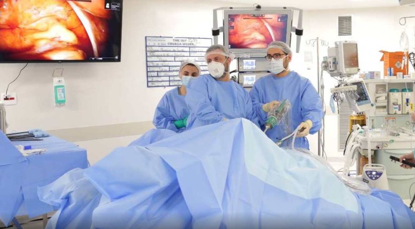 A foto mostra uma cirurgia robótica realizada pelos médicos do hospital. ês profissionais da saúde — dois homens e uma mulher — estão vestidos com aventais cirúrgicos azuis, toucas e máscaras, seguindo os protocolos de assepsia. Eles estão posicionados ao redor de um paciente coberto por campos cirúrgicos estéreis também azuis. O rosto do paciente não é visível. Dois dos profissionais (à esquerda e ao centro) observam o procedimento atentamente, enquanto o terceiro (à direita) manipula um aparelho com tubos, provavelmente um insuflador para cirurgia laparoscópica. A presença de dois monitores exibe imagens do interior do corpo do paciente em tempo real, sugerindo que se trata de uma videolaparoscopia, técnica minimamente invasiva. Ao fundo, há diversos equipamentos hospitalares, incluindo monitores de sinais vitais, um respirador, suportes para soro, e prateleiras com suprimentos médicos. Um quadro branco com o título “Time Out – Cirurgia Segura” está fixado na parede, reforçando os protocolos de segurança cirúrgica.