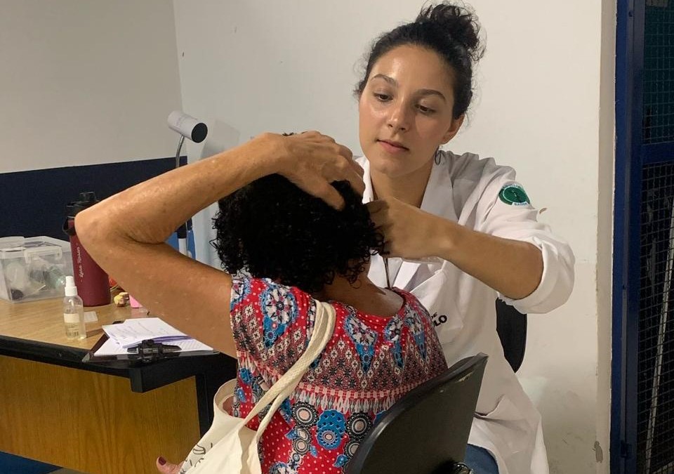 A imagem mostra uma profissional da área da saúde realizando atendimento a uma mulher. A profissional está usando um jaleco branco e parece estar examinando a orelha ou a cabeça da paciente, que está sentada de costas, com as mãos levantadas para cima da cabeça, facilitando o acesso da examinadora.  A paciente veste uma blusa estampada com padrões coloridos e carrega uma bolsa de tecido no ombro. O ambiente parece ser uma sala de atendimento simples, com uma mesa ao fundo contendo alguns materiais, como papéis, garrafa térmica, frascos e caixas organizadoras. A parede ao fundo é branca e azul escuro, e há um refletor ou abajur preso à mesa.  A cena transmite um momento de cuidado e atenção em um contexto de saúde