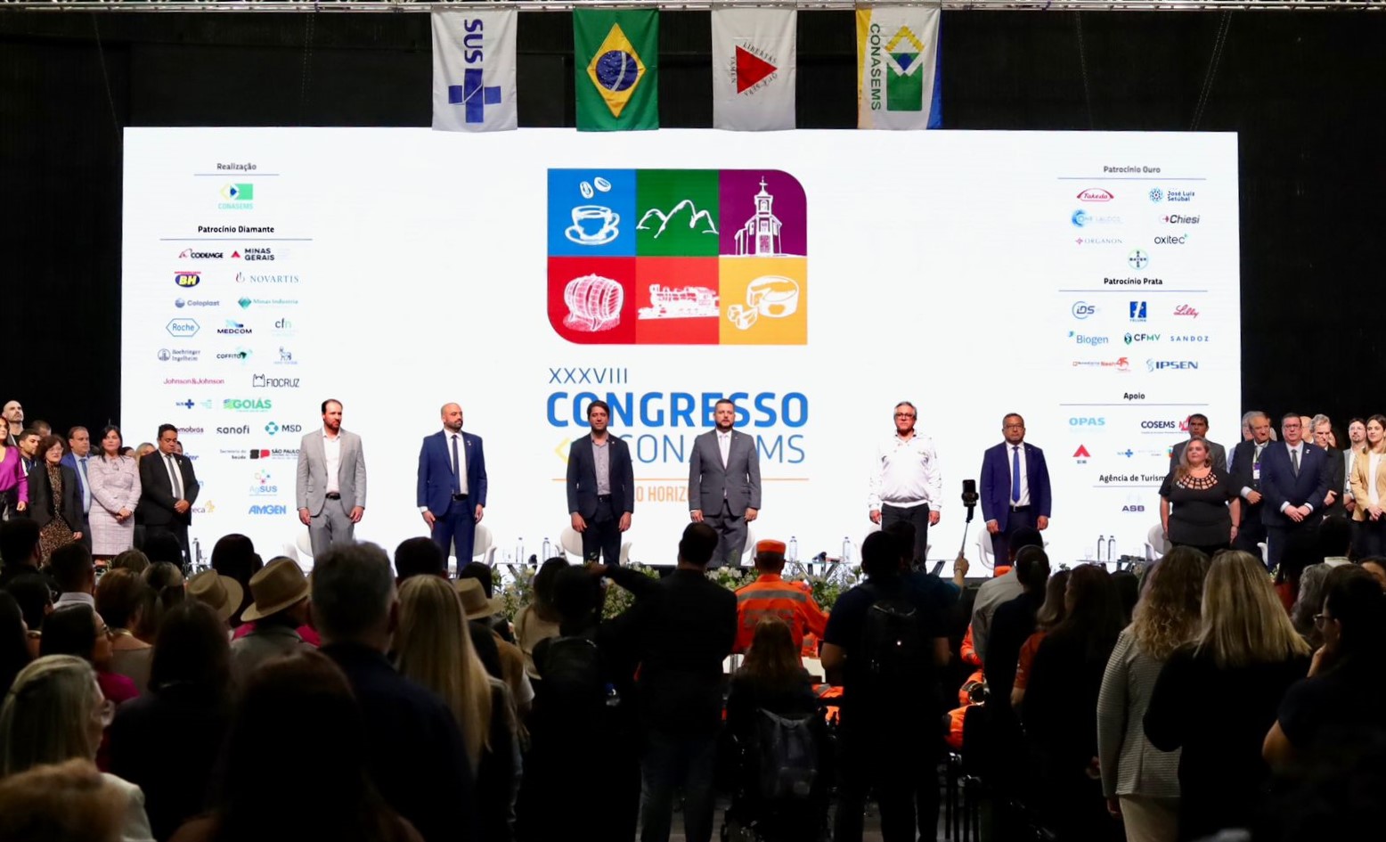 A imagem mostra o palco de um evento formal, com várias pessoas alinhadas em pé, formando uma fileira. Ao fundo, há um grande painel branco com o logotipo e o nome do evento: XXXVIII Congresso Conasems, indicando que é o 38º Congresso do Conselho Nacional de Secretarias Municipais de Saúde (Conasems), realizado em Belo Horizonte. O painel também exibe os logotipos de diversos patrocinadores e apoiadores. Acima do palco, estão hasteadas bandeiras, incluindo a do Brasil, a do SUS, a do Conasems e bandeiras de outros estados ou instituições. Na plateia, observa-se um público numeroso, com muitas pessoas em pé, algumas usando chapéus e outras com coletes laranja (possivelmente agentes de saúde ou defesa civil). Há também profissionais da imprensa e pessoas registrando o momento com celulares e câmeras. O clima geral é de solenidade e atenção.