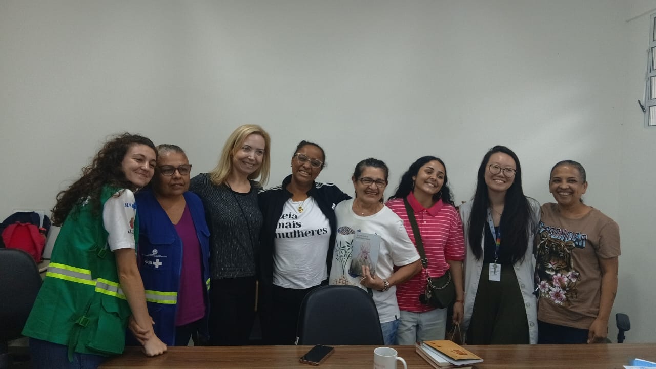 A imagem mostra um grupo de mulheres reunidas em uma sala simples, com parede branca e mesa de madeira à frente, dentro da UBS Parque Araribá. Ao centro está a escritora Lilia Guerra, que sorri enquanto posa junto às participantes. Ela está rodeada pelas idealizadoras e integrantes do clube de leitura da UBS Parque Araribá, formando uma composição afetiva e coletiva. Algumas mulheres usam crachás e vestimentas que remetem ao trabalho na saúde, enquanto outras estão com roupas casuais. Uma delas segura um livro, reforçando o contexto literário do encontro. Sobre a mesa, há objetos como um celular, uma caneca e alguns livros empilhados, indicando um momento de troca e conversa. A cena transmite proximidade, protagonismo feminino e valorização da leitura como prática de cuidado e convivência dentro do espaço da unidade de saúde.